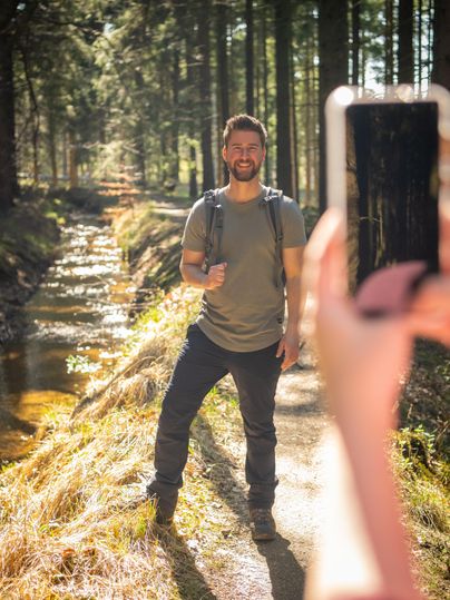 Ein lächelnder Mann steht auf einem Waldweg neben einem kleinen Bach. Er hält ein Handy in der Hand und posiert für ein Foto.