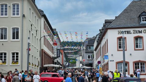Eine belebte Straße mit vielen Menschen und bunten Wimpeln. Auf beiden Seiten stehen historische Gebäude und Tische von Cafés sind aufgestellt.