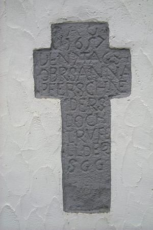 A gray stone cross with an engraved inscription. The cross is mounted on a white wall.