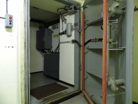 Ein Raum mit technischen Geräten und einem offenen Zugang. An den Wänden sind Rohre und Kabel sichtbar.