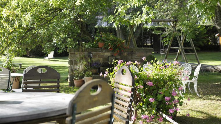 Ein einladender Garten mitHolztischen und -stühlen. Im Hintergrund sind grüne Bäume und eine Schaukel zu sehen.