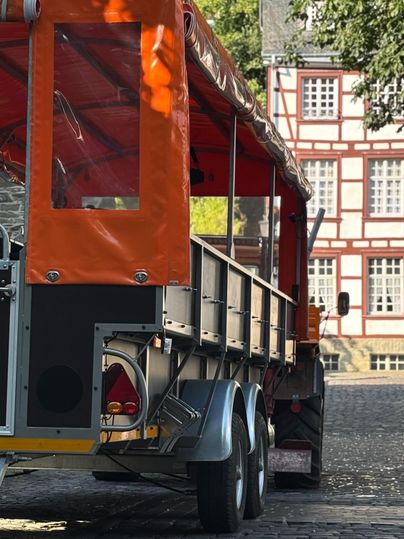 Ein orangefarbener Anhänger steht auf einer gepflasterten Straße. Im Hintergrund sind historische Gebäude zu sehen.