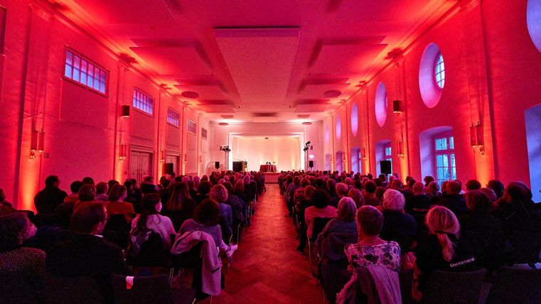 Ein großer Saal voller Menschen, beleuchtet in rotes Licht. Die Zuschauer sitzen in Reihen und blicken zur Bühne.