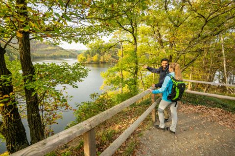 Ein Paar steht an einem Aussichtspunkt mit Blick auf einen See, umgeben von buntem Herbstlaub. Die Atmosphäre ist freundlich und entspannt.