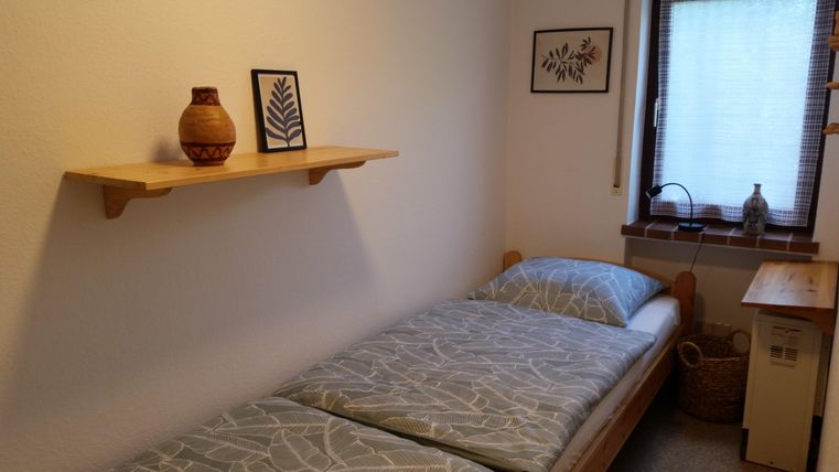 Ein einfaches Schlafzimmer mit einem Einzelbett und zwei Kissen. An der Wand hängt ein Regal mit einer Vase und zwei Bildern.