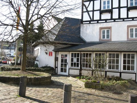 Ein charmantes Gebäude im Fachwerkstil mit großen Fenstern. Vor dem Haus befindet sich ein kleiner Platz mit Bäumen und einem gepflasterten Weg.