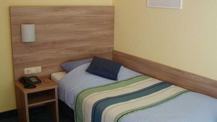 Een eenvoudige kamer met een eenpersoonsbed en een neutrale kleurstelling. Naast het bed staat een nachttafel met een telefoon.