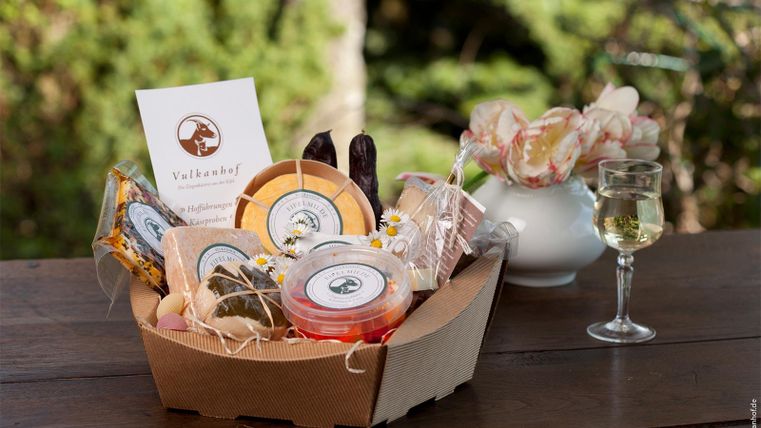 Ein Geschenkkorb mit verschiedenen Köstlichkeiten, darunter Käse und regionale Produkte. Daneben steht ein Glas Weißwein auf einem Tisch im Freien.