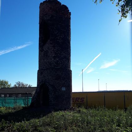 Een oude, gebroken toren van steen staat in een groene wei. Op de achtergrond zijn een blauwe lucht en enkele wolken te zien.