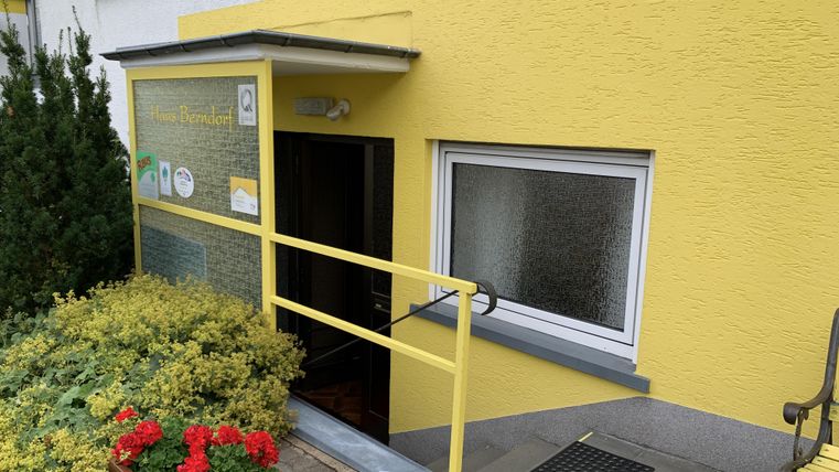 Eingang zu einem Gebäude mit gelber Fassade und einer Rampe. Vor der Tür blühen rote Blumen.