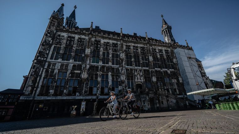 Eine alte, beeindruckende Fassade mit Türmen und vielen Fenstern. Zwei Radfahrer fahren in der Nähe des Gebäudes.
