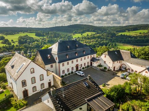Een schilderachtig landhuis omringd door groene heuvels en bomen. De lucht is helder met enkele wolken.