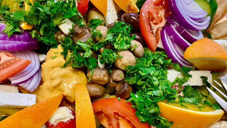 A colorful salad with fresh vegetables, herbs, and various fruits. Feta and pieces of bread are served with it.