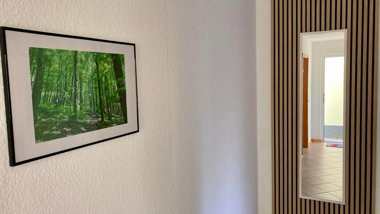 Ein heller Flur mit einer grünen Waldlanschaft in einem Bilderrahmen. An der Wand befindet sich ein vertikaler Spiegel mit einem gestreiften Rahmen.