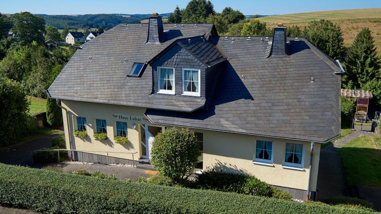 Een mooi huis met een steil dak en grote ramen. Omgeven door groene struiken en een landelijke omgeving.