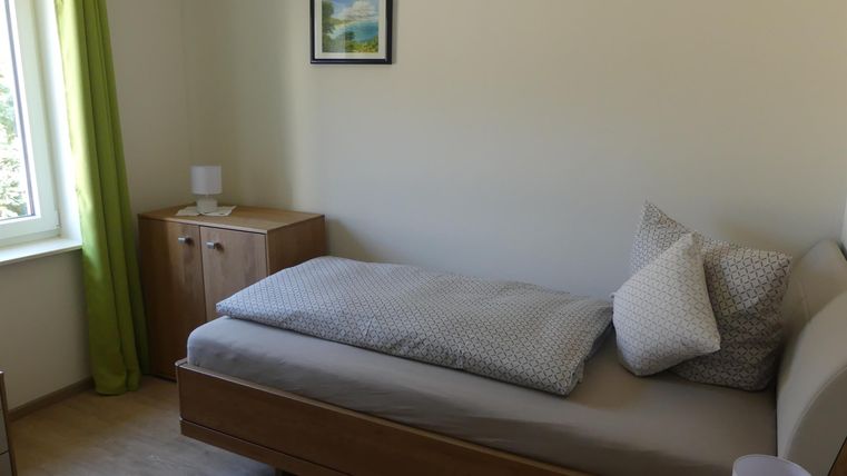 Ein schlichtes Schlafzimmer mit einem Einzelbett und einem Kissen. An der Wand hängt ein Bild, und die Fenster sind mit grünen Vorhängen dekoriert.