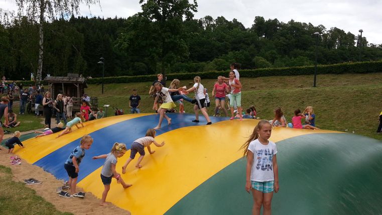Children are playing on a large, colorful inflatable cushion outdoors. Many people are gathered around the cushion. Trees are visible in the background.