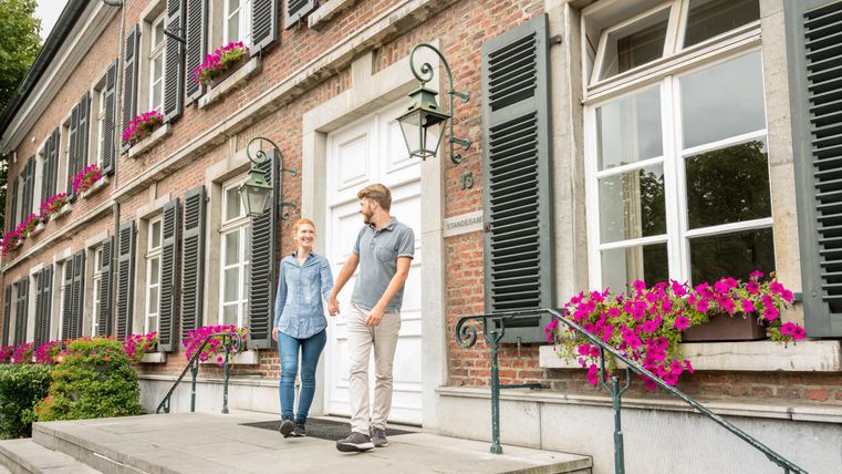 Een charmant gebouw met vensters die zijn versierd met groene luiken en kleurrijke bloemen. Twee personen staan op de trap en glimlachen.