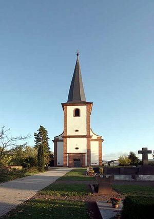 Eine Kirche mit einem hohen, spitzen Turm steht vor einem klaren Himmel. Im Vordergrund sind Gräber und eine gepflegte Grünfläche zu sehen.