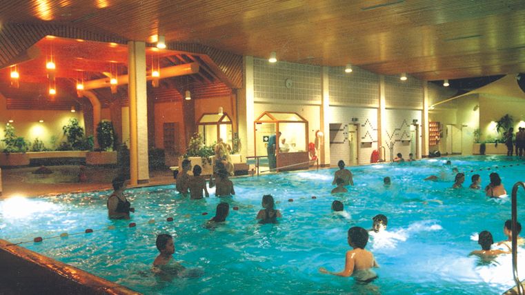 Een binnenzwembad met veel mensen die in het water zwemmen en spelen. De omgeving is helder verlicht en heeft een ontspannen sfeer.