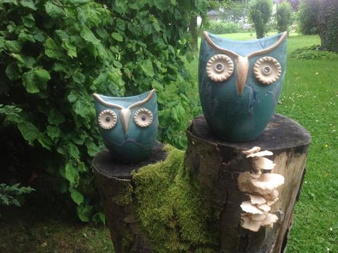 Twee decoratieve uilen van keramiek staan op een boomstronk. De achtergrond bestaat uit groene planten en gras.