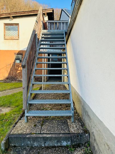 Eine Metalltreppe führt zu einem oberen Eingang. Im Hintergrund sind mehrere Gebäude und ein grüner Rasen zu sehen.