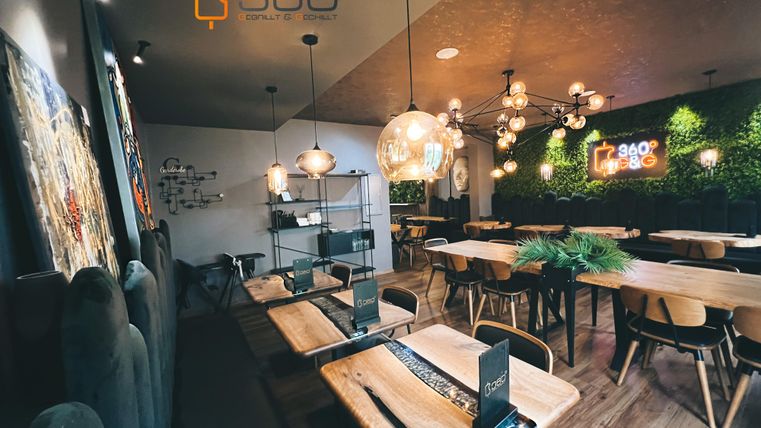 Een modern café met houten tafels en stijlvolle verlichting. De ontspannen sfeer wordt aangevuld met groene planten en een aantrekkelijke wanddecoratie.