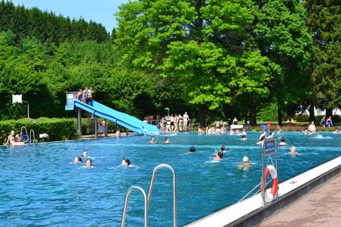 A swimming pool with clear water and many people swimming and relaxing. In the background, there is a slide and lush greenery.