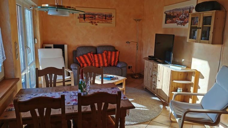 Een gezellige woonkamer met een eettafel en stoelen. Op de bank liggen oranje kussens en een televisie staat op een houten kast.