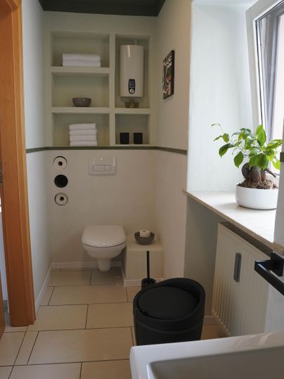 Ein modernes Badezimmer mit einer Toilette und einem Waschbecken. Es gibt Regale mit Handtüchern und eine Pflanze auf der Fensterbank.