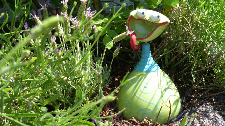 Een kleurrijke tuinfiguur in de vorm van een slang staat tussen groen gras en kleurrijke bloemen. Ze heeft een vrolijk gezicht en houdt iets in de hand.