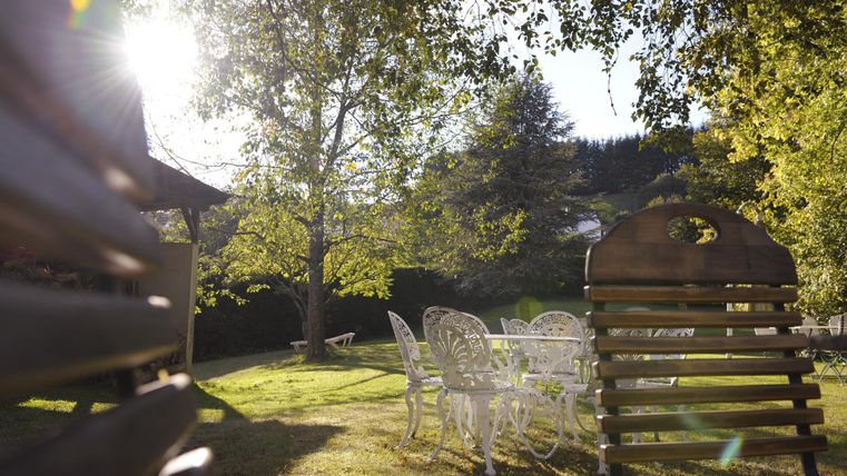 Ein sonniger Garten mit einem Tisch und Stühlen aus Weiß. Im Hintergrund stehen grüne Bäume und die Sonne scheint durch die Blätter.