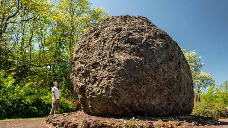 Ein großer, runder Fels steht in der Natur. Neben dem Felsen steht eine Person, die ihn betrachtet.