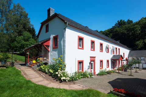 Ein charmantes, weißes Haus mit roten Fensterrahmen und einem schönen Garten. Im Hintergrund sind Bäume und blauer Himmel zu sehen.