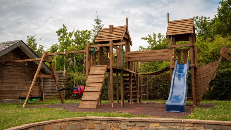 Een houten speeltuin met glijbanen, ladders en schommels. De omgeving is groen en uitnodigend.