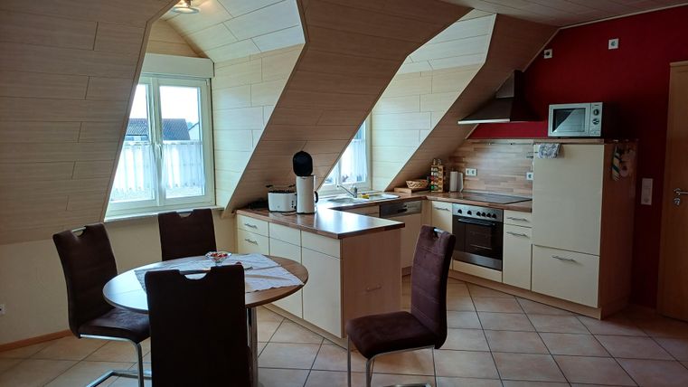 A modern attic kitchen with slanted walls. The dining area consists of a round table and chairs, surrounded by window light.