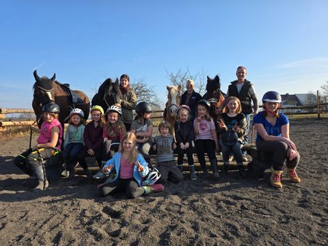 A group of children and adults stands and sits at a riding stable, surrounded by horses. The atmosphere is cheerful and sunny.