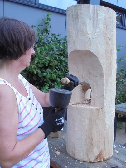 A woman is carving on a wooden block outdoors. She is using a tool to create an opening in the wood.
