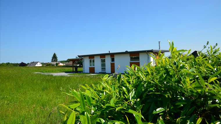 A modern house stands in the middle of a green meadow under a clear blue sky. In the background, more houses are visible.