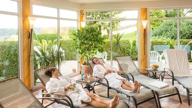 Entspannte Menschen in Bademänteln liegen auf Liegestühlen in einem hellen Raum. Große Fenster lassen viel Tageslicht und Blick ins Grüne herein.