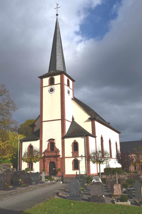 Eine Kirche mit einem hohen spitzen Turm und einem klaren Wetterhimmel. Im Vordergrund sind Gräber zu sehen.