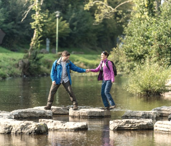 Een stel kruist stapstenen in de Kyll bij Gerolstein. Ze houden elkaars hand vast en dragen outdoorkleding., © Eifel Tourismus GmbH, Dominik Ketz
