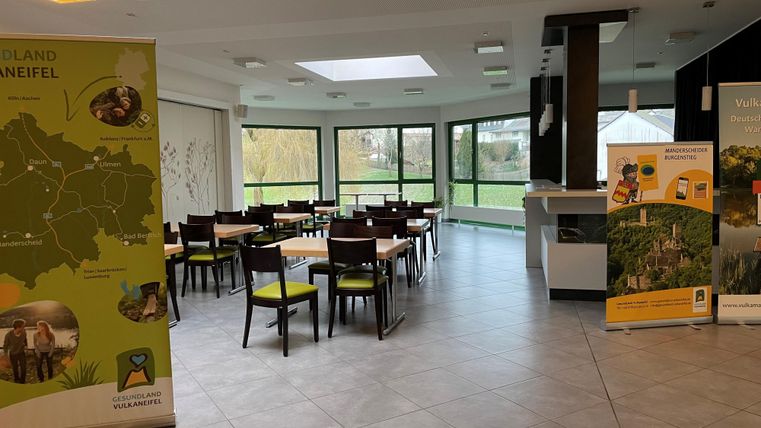 Ein einladender Raum mit Tischen und Stühlen sowie großen Fenstern, die auf einen grünen Außenbereich blicken. An den Wänden sind Informationsbanner angebracht.