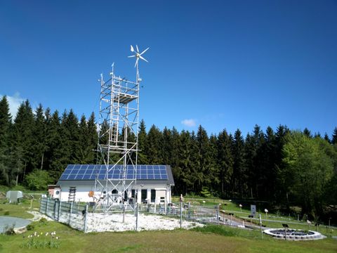 Ein Gebäude mit Solarpanelen und einem Windrad in einer ländlichen Umgebung. Der Himmel ist klar und blau, und Bäume umgeben das Areal.