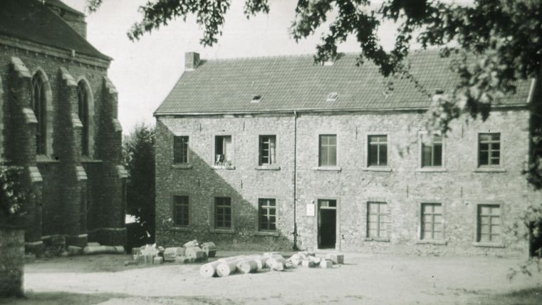 Een oud stenen gebouw met veel ramen staat op een hof. Enkele zakken liggen op de grond en bomen werpen schaduw op de scène.