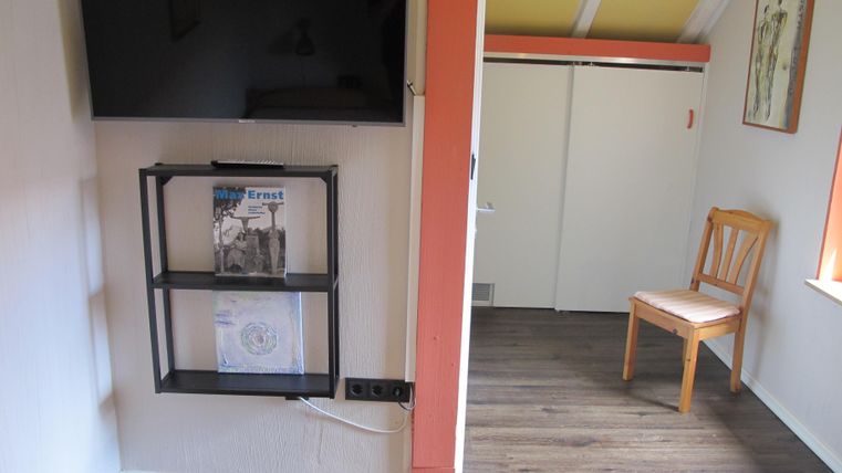 Ein gemütlicher Raum mit einem Fernseher an der Wand und einem Holzstuhl in der Ecke. Der Boden ist mit hellem Laminat ausgelegt.