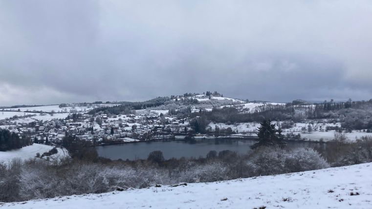 Eine winterliche Landschaft mit Schnee bedeckten Hügeln und einem ruhigen See. Im Hintergrund sind kleine Häuser und bewaldete Flächen zu sehen.