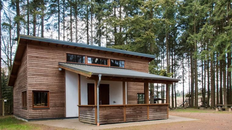 Ein modernes Holzhaus in einem Waldgebiet. Es hat eine Veranda und große Fenster, die viel Licht hereinlassen.