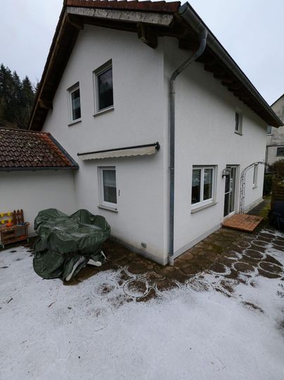 A two-story white building with a gabled roof. In the foreground is a paved area with a covered object.