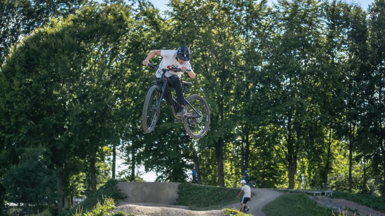 Ein BMX-Fahrer springt in der Luft über eine Sprungschanze. Im Hintergrund sind grüne Bäume zu sehen.